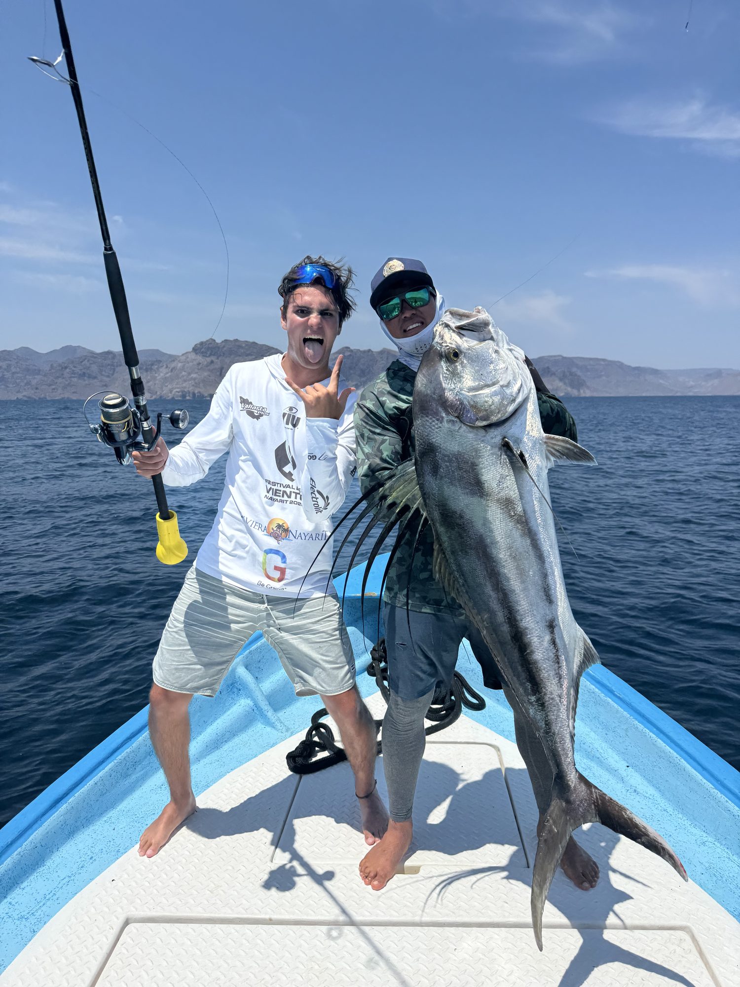 Santiago on a boat holding a fishing rod, next to a roosterfish held up by his guide